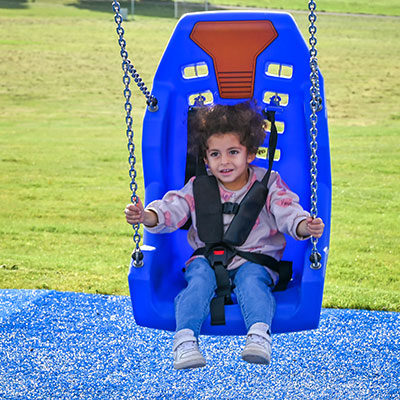 A little toddler girl is swinging on a mirage swingseat at a playground, holding on to the chains and smiling.